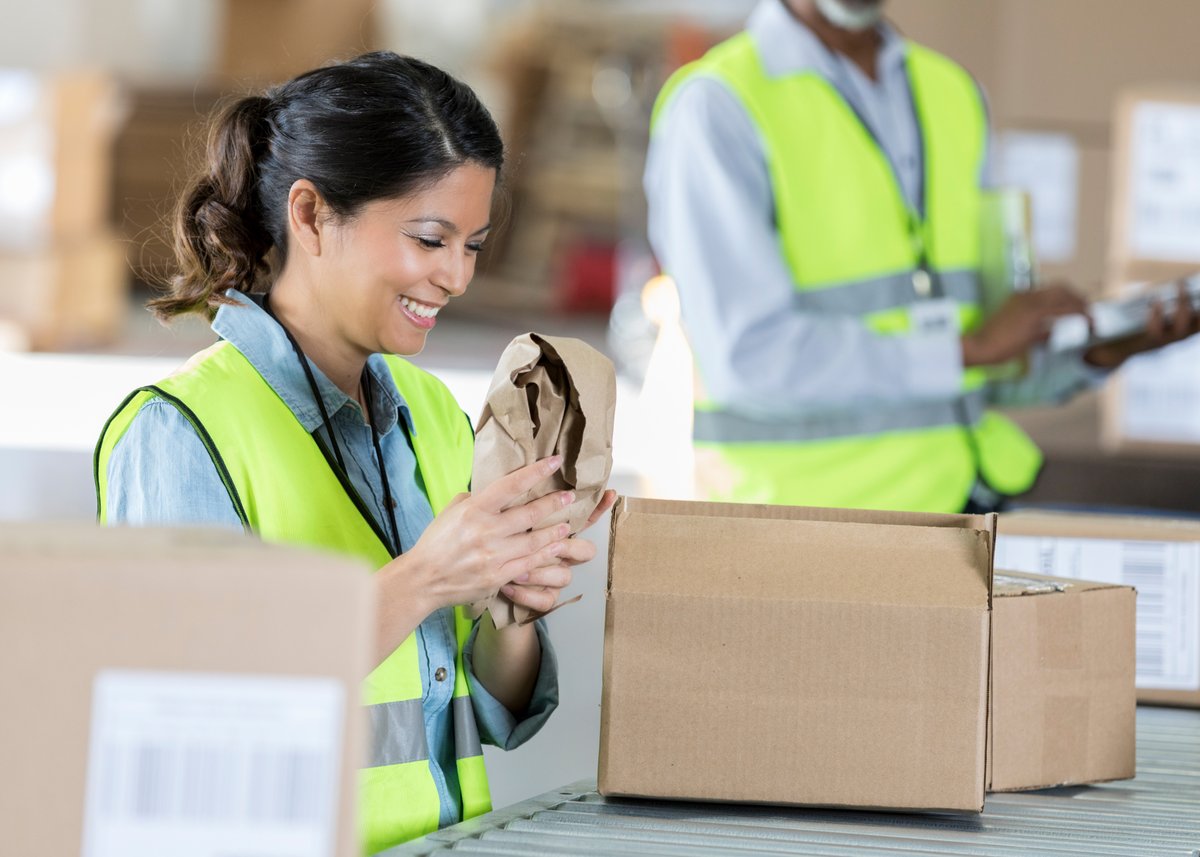 A female warehousing employee in a lime green vest repacks the customer's product. 